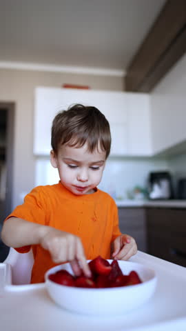 Cute Caucasian boy chooses a big red strawberry from the plate. Toddler kid eats healthily at home in the kitchen. Close up. Vertical video.