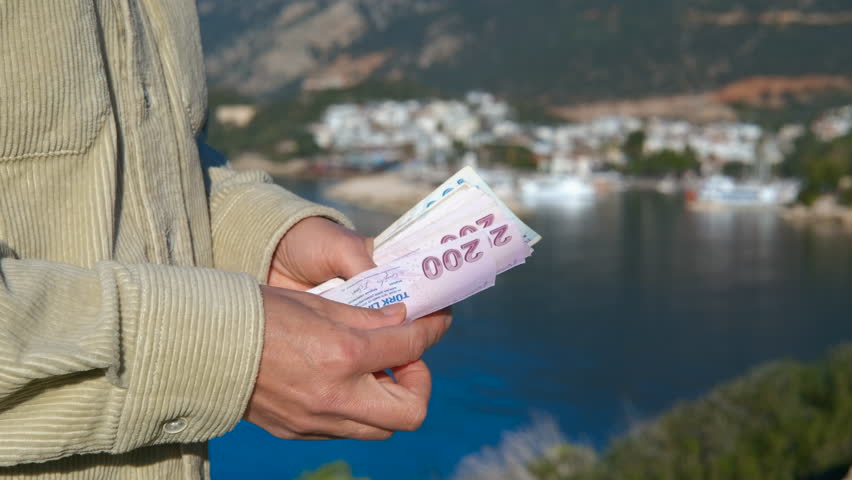 Tourist standing near coastal scene while carefully counting turkish lira banknotes against scenic seaside landscape with bright summer sky