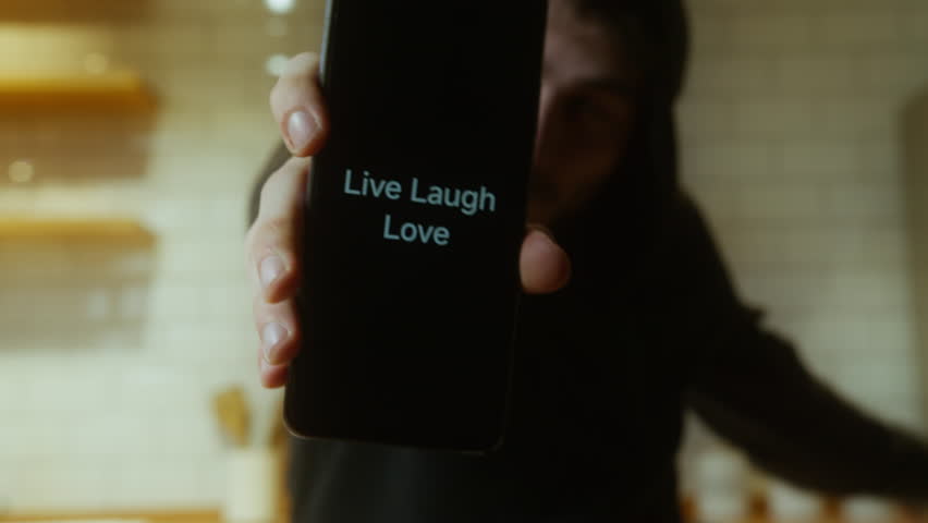 Cheerful Young Man Holding Smartphone Displaying Live Laugh Love Message in Bright Kitchen Environment