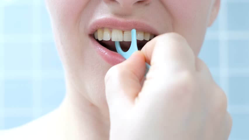 Young woman brushing teeth with disposable dental floss close-up against the background of blue tiles in the bathroom.