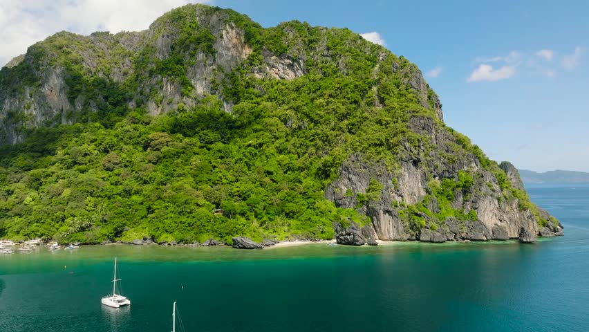 Pristine green hills on islands with rocky coast surrounded by vibrant turquoise waters with a sailboat. El Nido, Philippines.