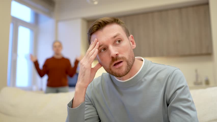 Frustrated exhausted man rolls eyes tired of constant fights, woman in background yelling waving hands. Accusations infidelity, marriage conflict, love faded, emotional strain, relationship fall apart