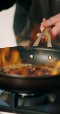 Hands, chef and flame with frying pan in kitchen for cooking with ingredients, meal prep and gourmet cuisine in home. Person, stove and fire with food, nutrition and seasoning with oil at apartment