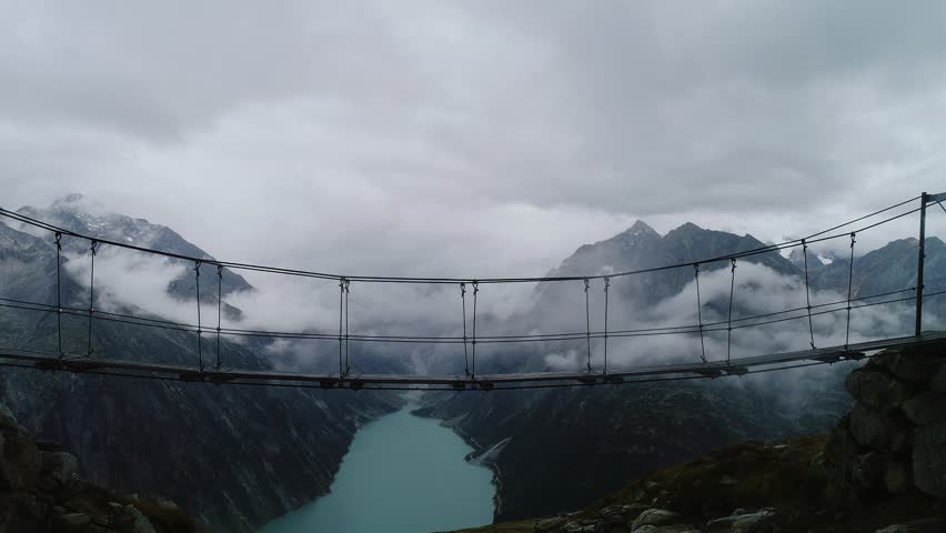 Hiker walks hanging bridge above turquoise lake and misty peaks in moody Austria