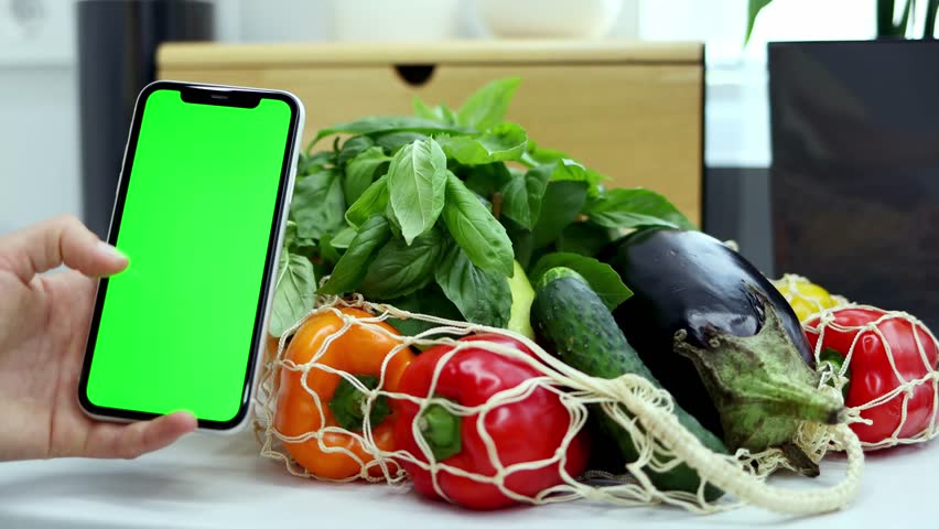 Close-up of female hands holding smartphone with green screen on a background of organic vegetables in the modern kitchen. Concept of online shopping and food delivery. Chroma key. 4k footage.