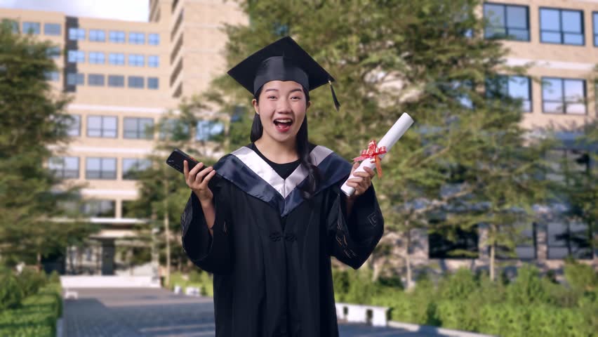 Asian Woman Student Graduates In Cap And Gown With Diploma Looking At Smartphone Then Saying Wow In Front Of A Magnificent University Building