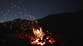 Night campfire, hike mountains. Black silhouette of mountains in background against sky. Burning logs, hearth. Sparks from fire explode like fireworks in dark. Fire magic. Amazing night by campfire - Powered by Shutterstock - Get 15% off with code: PIKWIZARD15