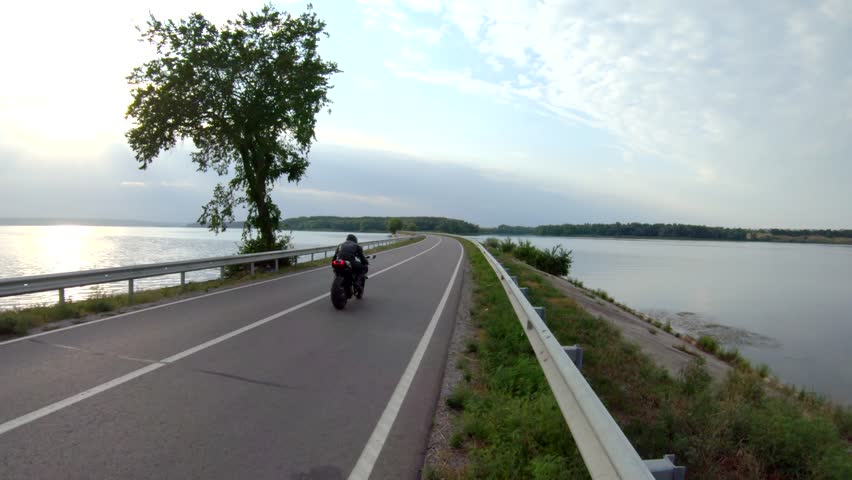 Biker riding on modern sport motorbike through road near lake. Motorcyclist racing motorcycle at dam route on summer day. Man driving bike through bridge of river. Concept of freedom and lifestyle