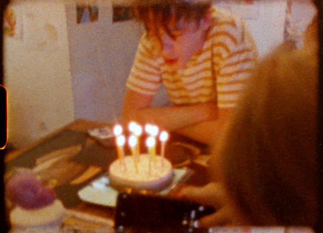 Young boy celebrating birthday with lit candles on cake, captured through nostalgic Super 8 film effect while family member records moment using smartphone
