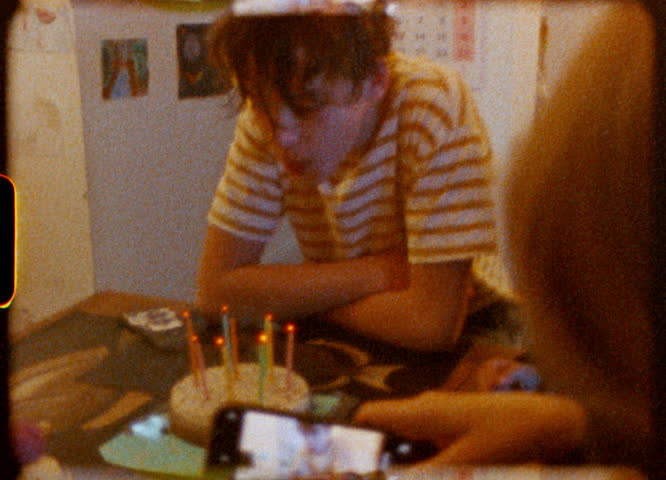 Young boy celebrating birthday with lit candles on cake, captured through nostalgic Super 8 film effect while family member records moment using smartphone