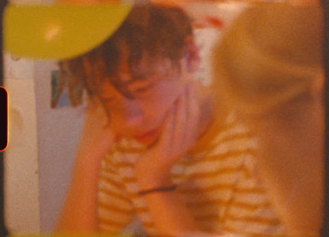 A quiet birthday moment captured on Super 8mm film. A teenage boy gazes dreamily at his birthday cake while his mother lights the candles. The soft glow of the flame creates a nostalgic, intimate