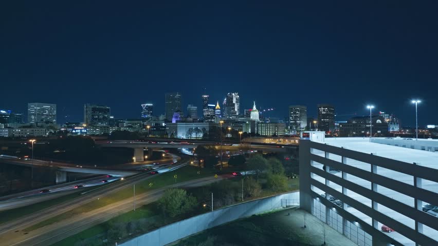 Car park with busy highway at night in Atlanta City, Georgia. Aerial forward wide shot. Skyline and skyscrapers in distance. Lighting buildings of downtown.