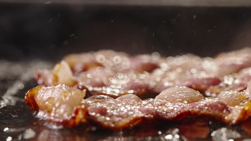 Frying pork bacon in pan side view close up. Aroma of bacon frying in pan. Fried to perfect. Sweet-savory taste of crispy bacon alongside morning breakfast. Cholesterol occasionally enjoying bacon