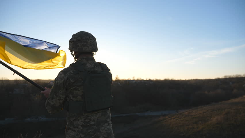 Ukrainian army soldier holding waving flag of Ukraine. Portrait of man in military uniform and helmet lifted up flag in hill. Victory against russian aggression. Invasion resistance concept. Slow mo