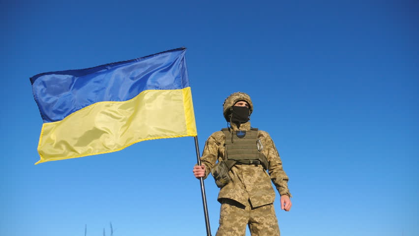 Ukrainian army man in military uniform lifting national banner. Young male soldier holds a waving flag of Ukraine against blue sky. Victory against russian aggression. Invasion resistance. Low view