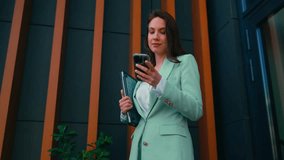 Young businesswoman in a light green suit walks outside an office building, using her smartphone for calls and messages while holding a laptop - Powered by Shutterstock - Get 15% off with code: PIKWIZARD15