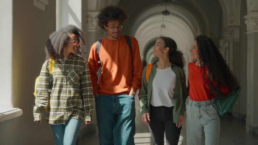 Group of happy diversity students walking in university corridor talking smiling four friends walk in high school hall discuss African American man guy with Arabian Hispanic women girls go to class