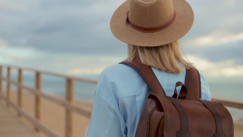 Blonde traveler with straw hat, leather backpack traversing wooden pier, soaking in coastal summer scenery.Travel, weekend, relax and lifestyle concept