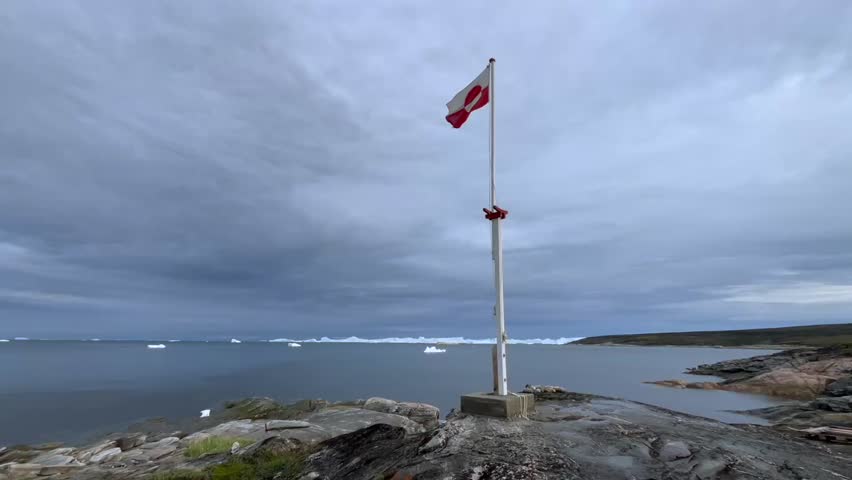 Greenland’s national flag fluttering on a coastal cliff near Ilulissat, with icebergs floating in the Arctic Ocean beneath a cloudy sky.