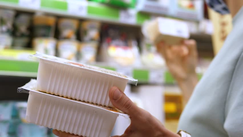 Focused female shopper holding tofu packages while reaching across refrigerated shelf, carefully selecting plant based protein product in grocery store environment