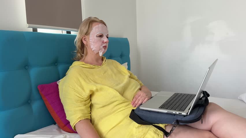 Woman use of paper mask at home woman with laptop working sitting on bed in bright yellow dress
