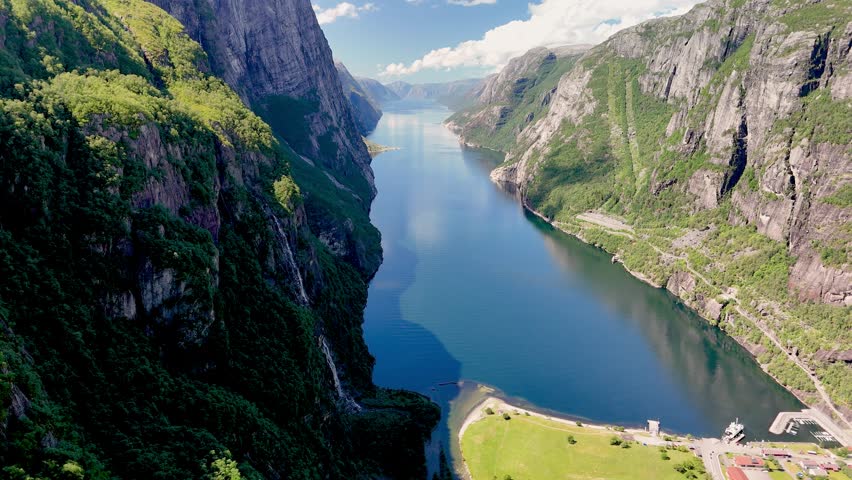 Majestic cliffs rise steeply along the tranquil waters of a Norwegian fjord. Lush greenery carpets the mountainsides, showcasing natures splendor. Lysebotn, Lysefjorden, Norway