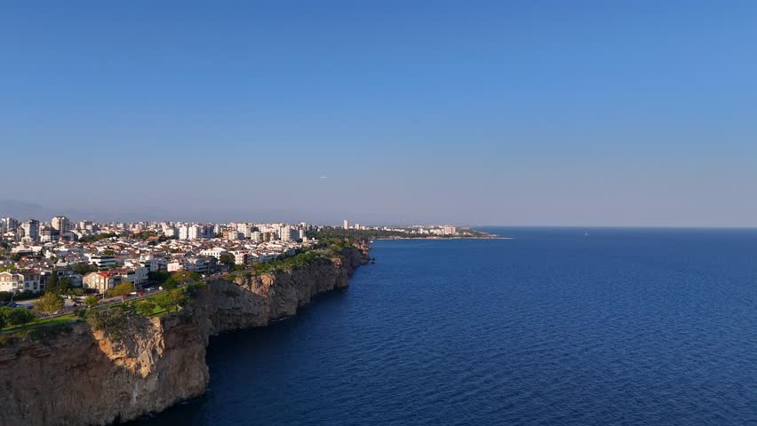 Still drone shot of seaside city and rocky cliffs beneath clear blue sky, featuring distant airplane over the ocean horizon.
