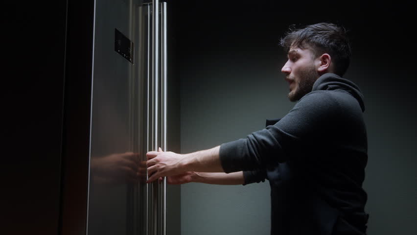 Young Man Holding a Jar and Looking Inside Refrigerator Late at Night in Dim Kitchen