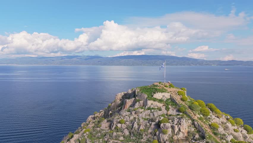 An orbit drone shot reveals the traditional port of Hydra as it circles around the Greek flag on the hilltop. A powerful blend of symbolism, island charm, and scenic beauty