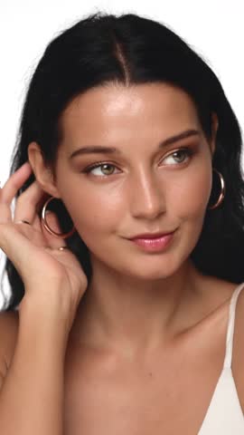 A stunning Latina woman with dark hair and hoop earrings gently touches her face, admiring her beauty and glowing skin. Ideal for skincare, makeup, self-care, and cosmetic branding concepts.