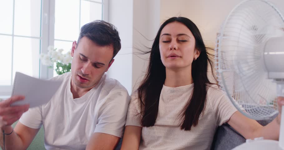 Family couple faces discomfort and trouble from hot summer weather at home living room, using fan for cool air in a challenging setting with focus on managing heat and relief every day.
