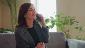 Concerned businesswoman in gray suit sitting on couch speaking on phone with serious look and hand gesture in bright indoor space with houseplants and soft daylight streaming through window - Powered by Shutterstock - Get 15% off with code: PIKWIZARD15
