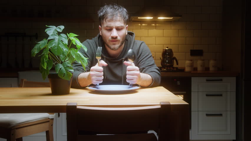 Hungry man sitting at kitchen table waiting for food with empty plate and cutlery in hand