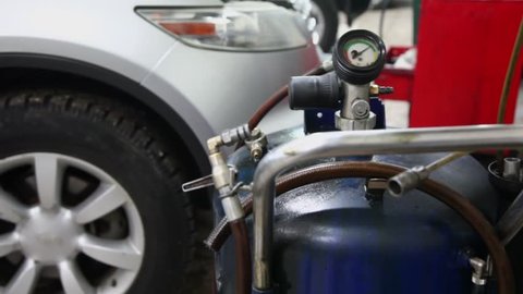 Gas cylinder with pressure gauge stands near car with open hood in automobile service station