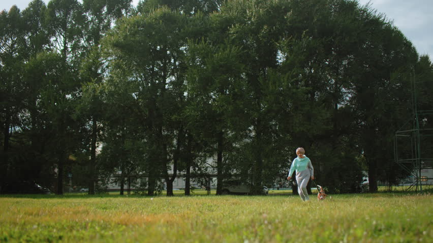 Wide view of trainer running with dog in lush green natural park during bright summer day, surrounded by tall trees, open grassy field, relaxed joyful outdoor atmosphere perfect for pet exercise