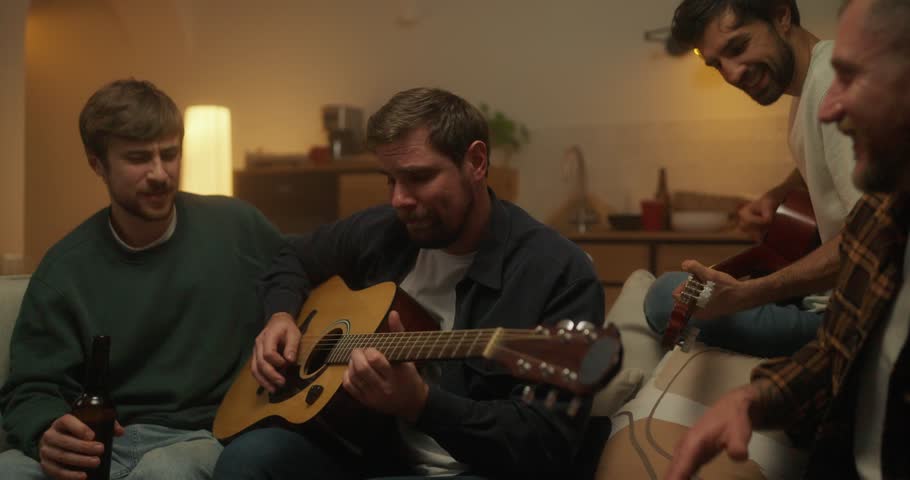 A brunette guy plays the guitar with his friend while their friends play along with them while relaxing at home at a bachelor party in the evening