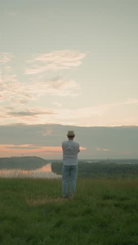 A gentleman stands contemplatively in a grassy field by a tranquil lake at sunset. Dressed in a white shirt, hat, and jeans, he captures a peaceful, reflective moment as the sun sets