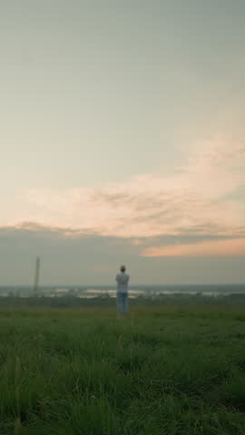 A man wearing a white shirt, hat, and jeans stands contemplatively in a grassy field by a tranquil lake at sunset. The serene landscape and warm evening light create a peaceful, reflective scene