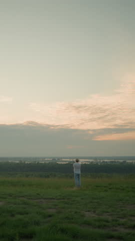 An adult male, in a white shirt, hat, and jeans, stands contemplatively in a grassy field by a tranquil lake at sunset. The serene landscape and warm light create a peaceful, reflective atmosphere