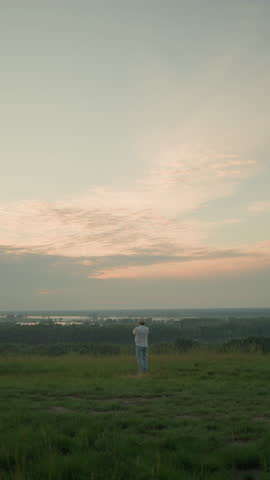 A distant view of a man dressed in a white shirt, hat, and jeans, standing contemplatively in a grassy field by a tranquil lake at sunset. The expansive sky and serene setting evoke a sense of peace
