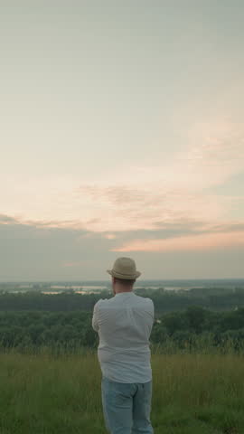 A thoughtful man wearing a white shirt, hat, and jeans stands in a grassy field by a tranquil lake at sunset, gazing into the distance. The warm sunset light create a peaceful atmosphere