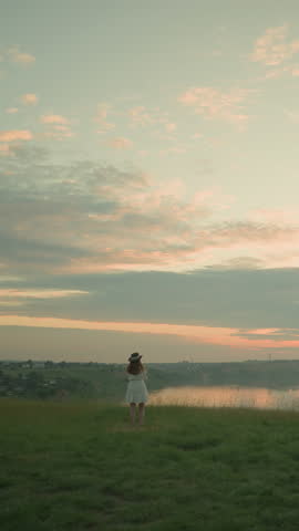 Back view of a woman in a white dress and hat standing alone in an open grassy field, gazing at a calm lake and distant village during a serene sunset, under a cloudy, pastel-colored sky