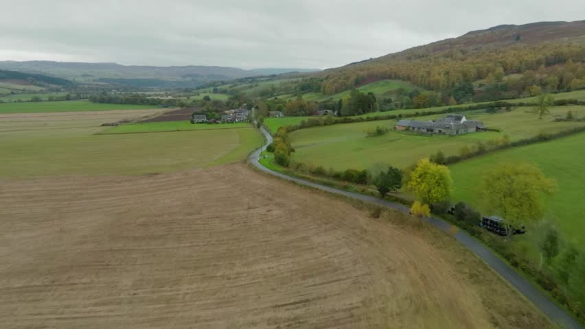 Car driving along countryside road through farmland in Scottish Highland valley and rolling hills, aerial shot over scenic route