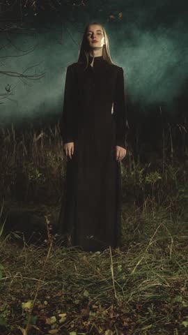 Woman with long blonde hair is posing in a dark forest. She is wearing a long black dress and the atmosphere is eerie and mysterious