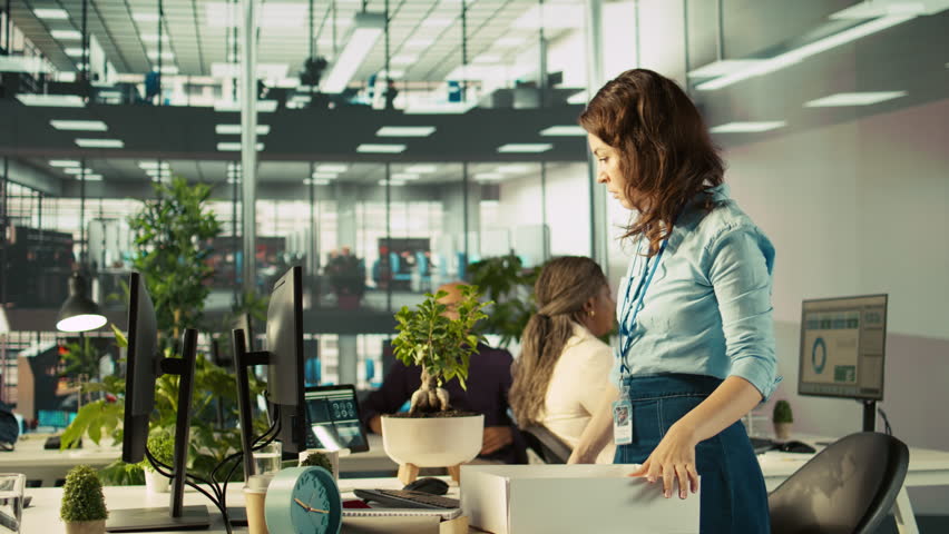 Upset worker in office packing desk plants on last day at work after quitting, saying goodbye to colleague. Employee getting fired from job during layoffs, saddening coworkers, camera B