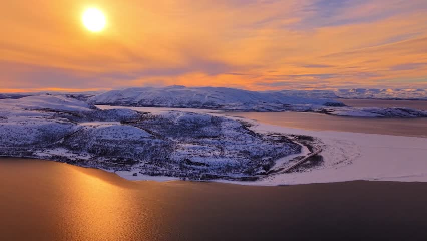 Gorgeous aerial view of white snow and ice covered Giemaš mountain that is surronded by ocean water Barents sea during a golden sunrise or sunset in nordic Norway. Calming scene with horizon visible.