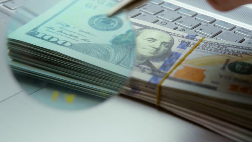 Close-Up of Counterfeit Money Inspection with Ultraviolet Light on Dollar Bills Over Laptop