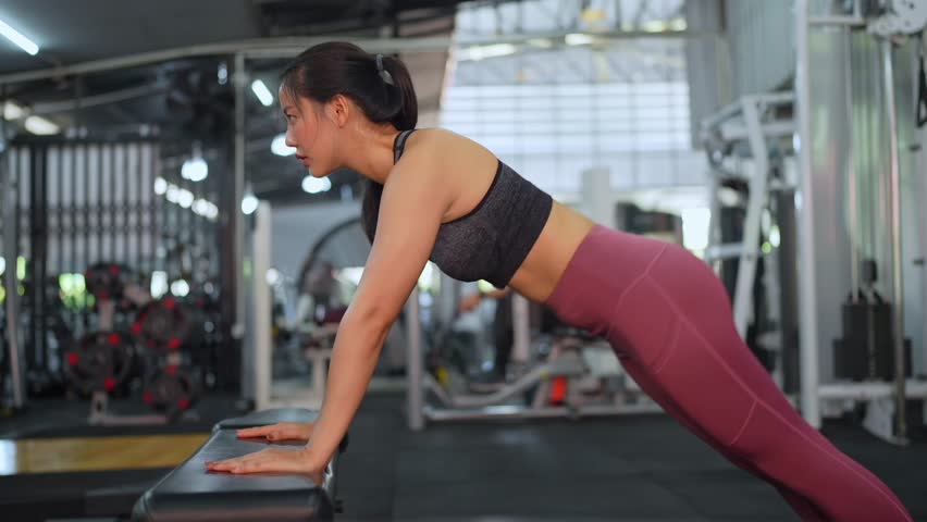 Young asian sportswoman wearing sportswear performing incline push ups exercise using bench in fitness gym, demonstrating strength and dedication to fitness goals