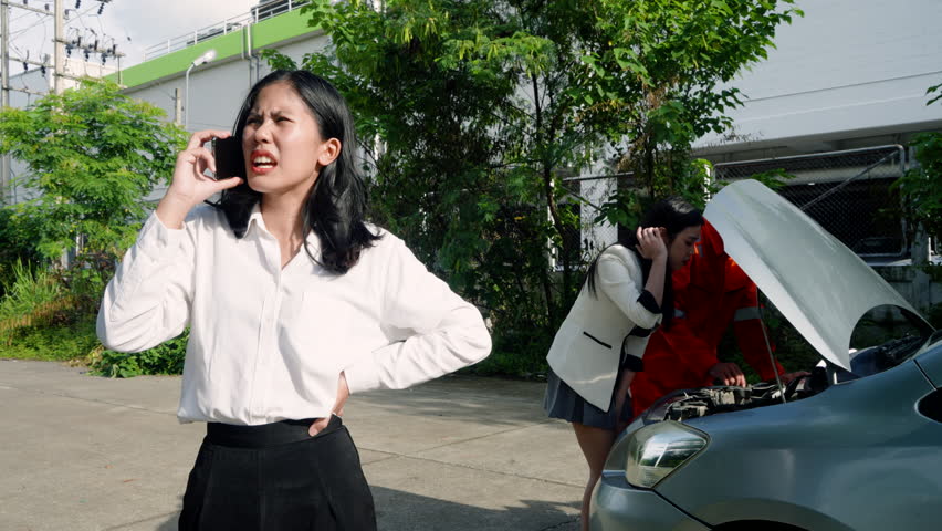Asian adult female customer making phone call standing beside broken-down car while insurance agent and another woman inspect engine during roadside emergency in outdoor urban area