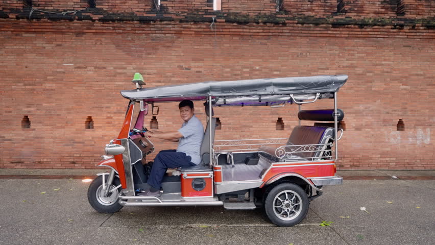 Asian adult male tuk tuk driver wearing short sleeve shirt and long pants sitting in red motorized rickshaw smiling parked on street with ancient brick wall in background in Chiang Mai Thailand